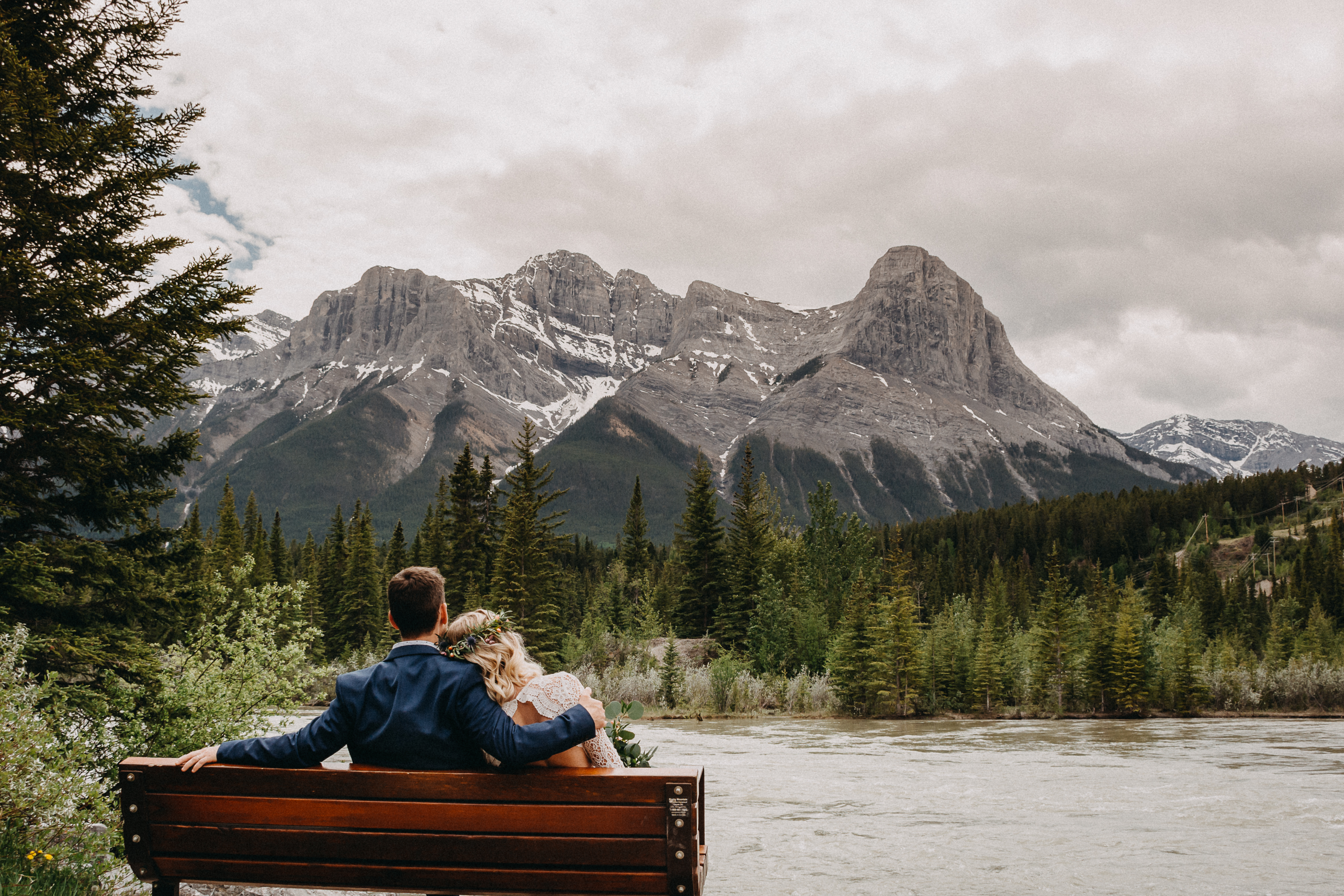 Groom and Bride sitting on bench in front of a river and a section of the rocky mountains
