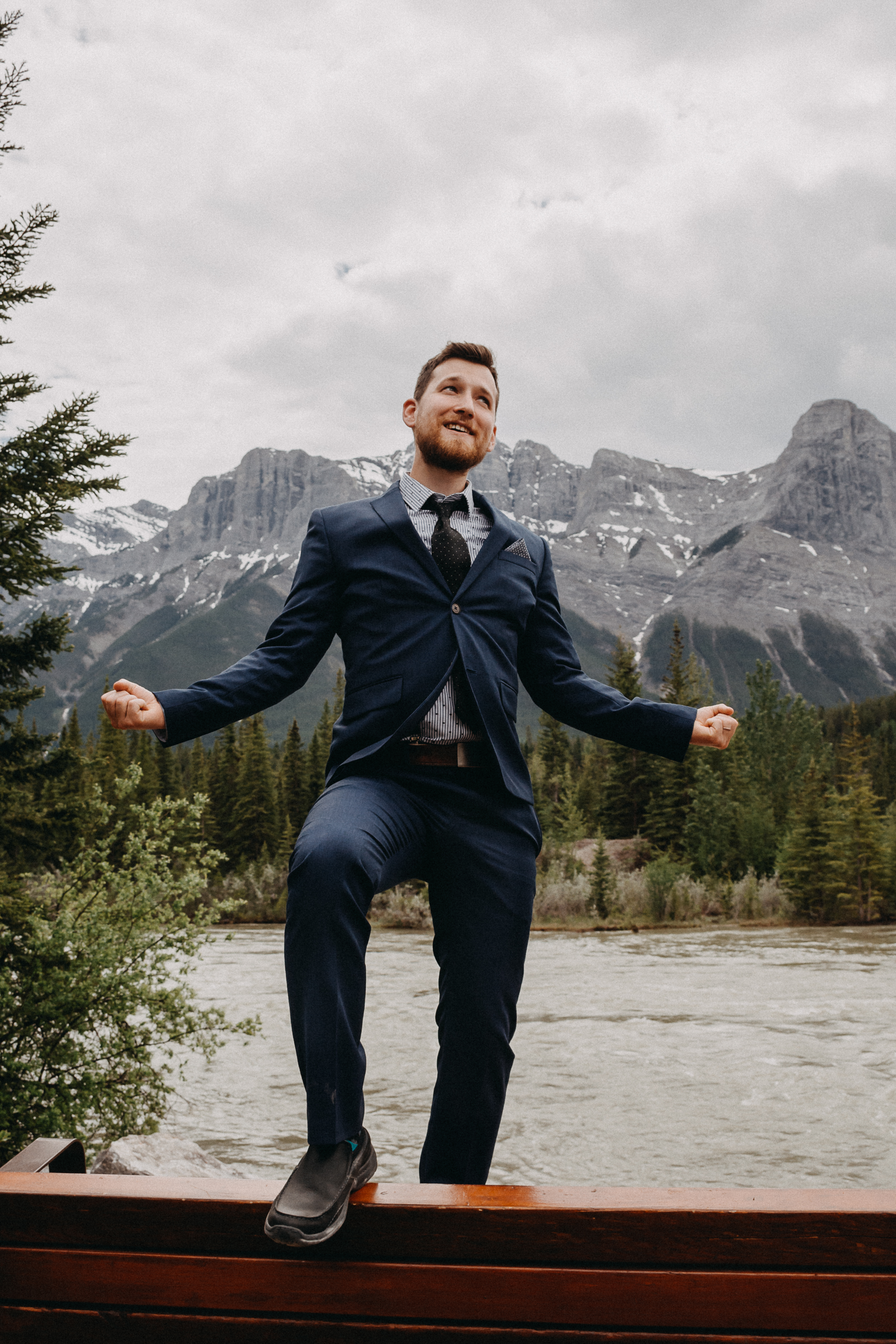 The groom standing on top of a bench in front of the rocky mountains, expressing joy
