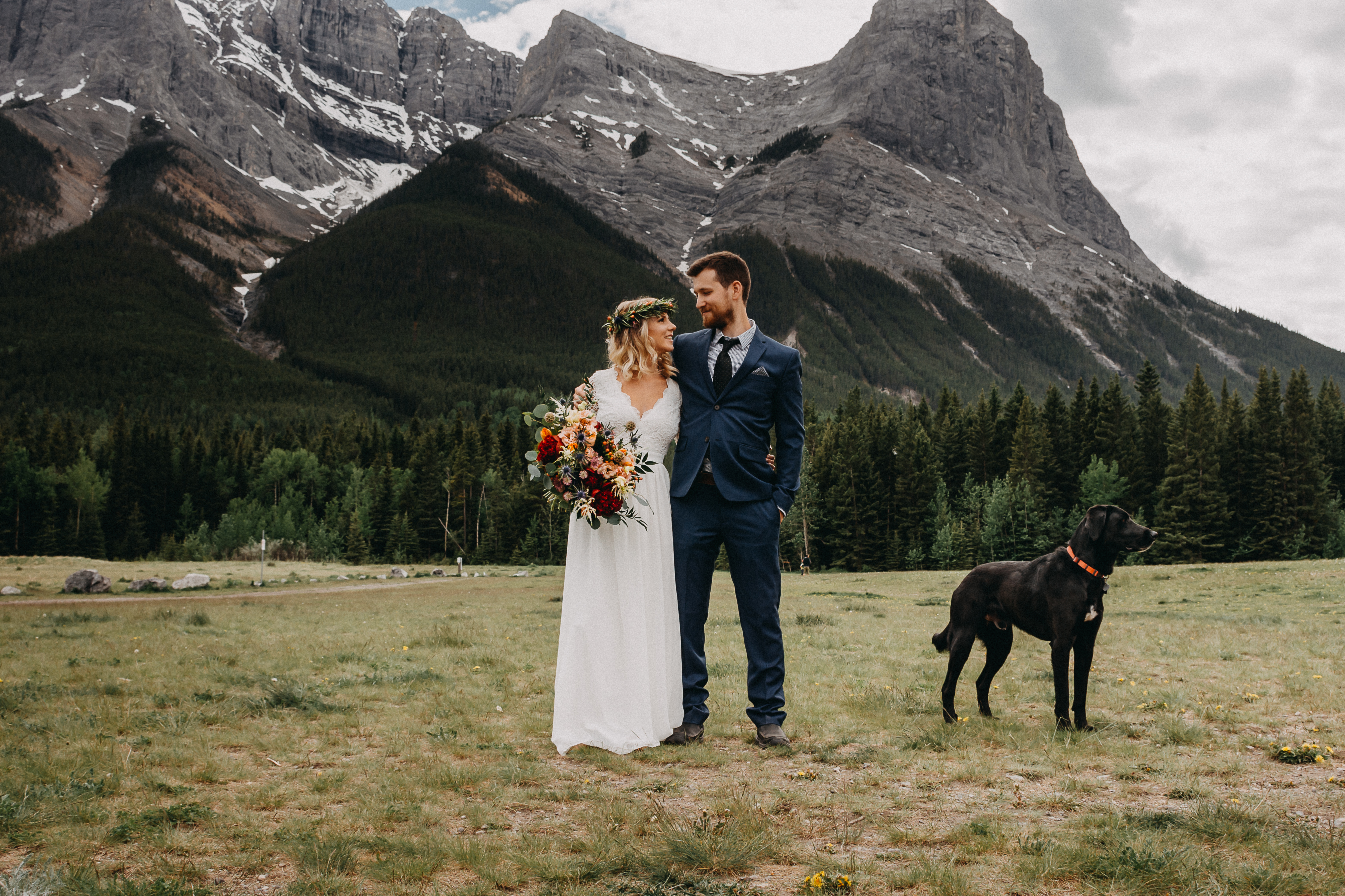 The bride and groom looking at each other and their dog in front of the rocky mountains