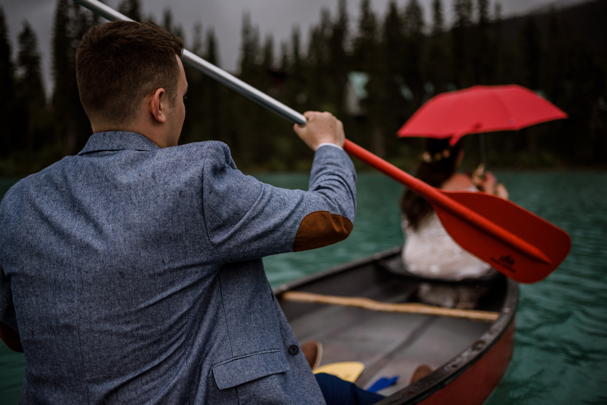 Close up groom paddling a canoe with bride holding red umbrella at Emerald Lake, BC
