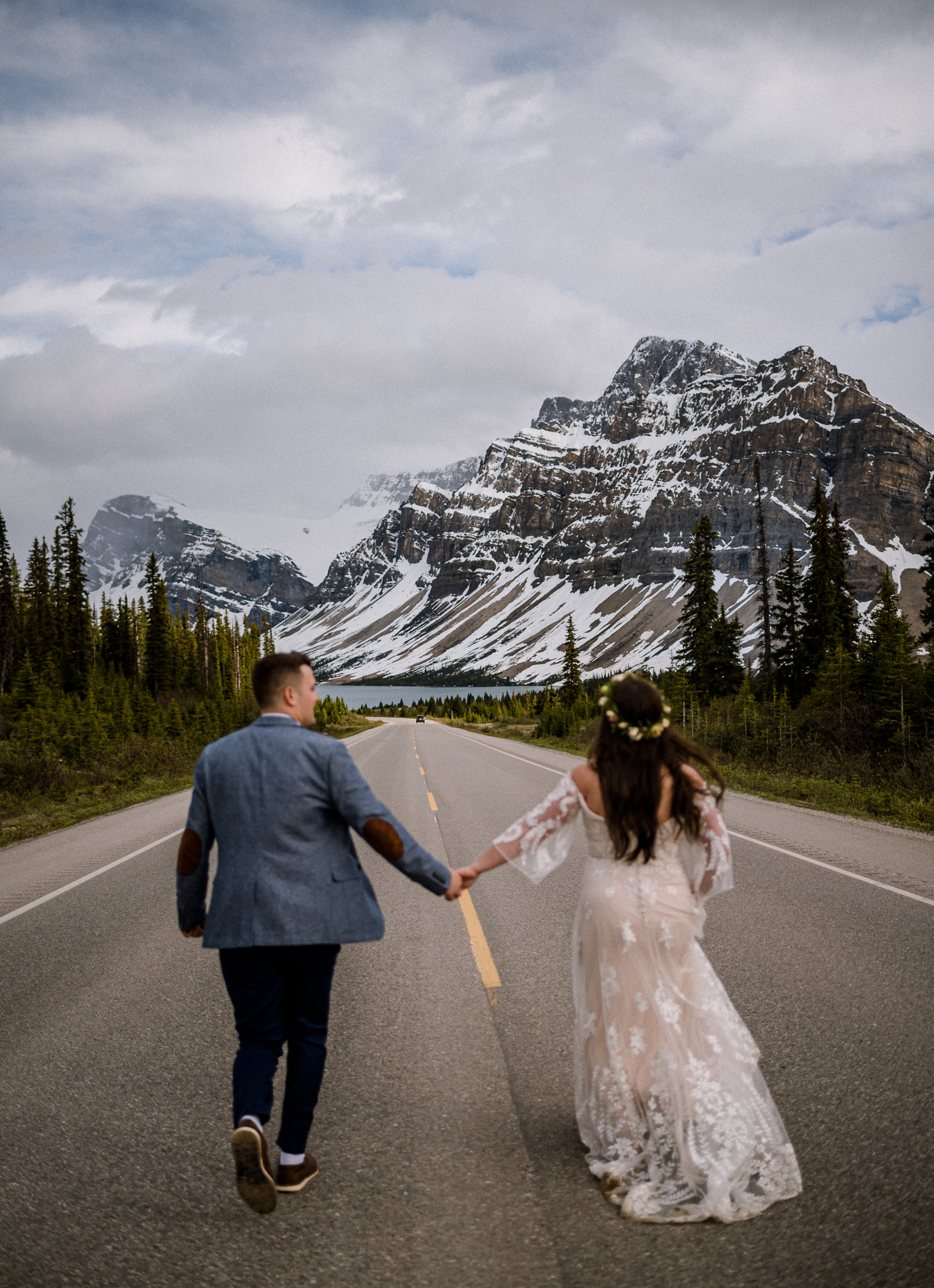 Groom and bride holding hands running down empty road in Alberta with mountains in focus