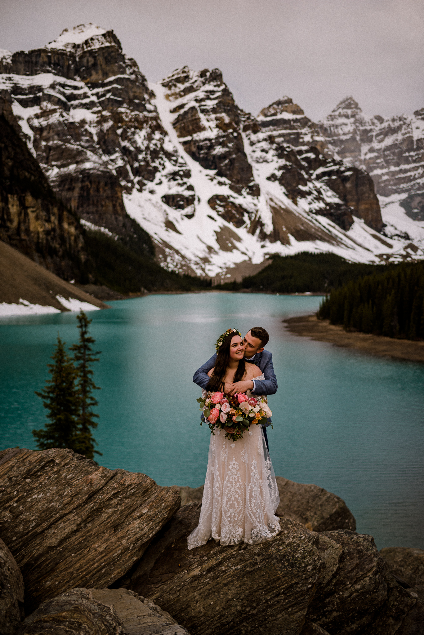 Groom hugging bride from behind while she holds bouquet and laughs in front of Moraine Lake, Alberta