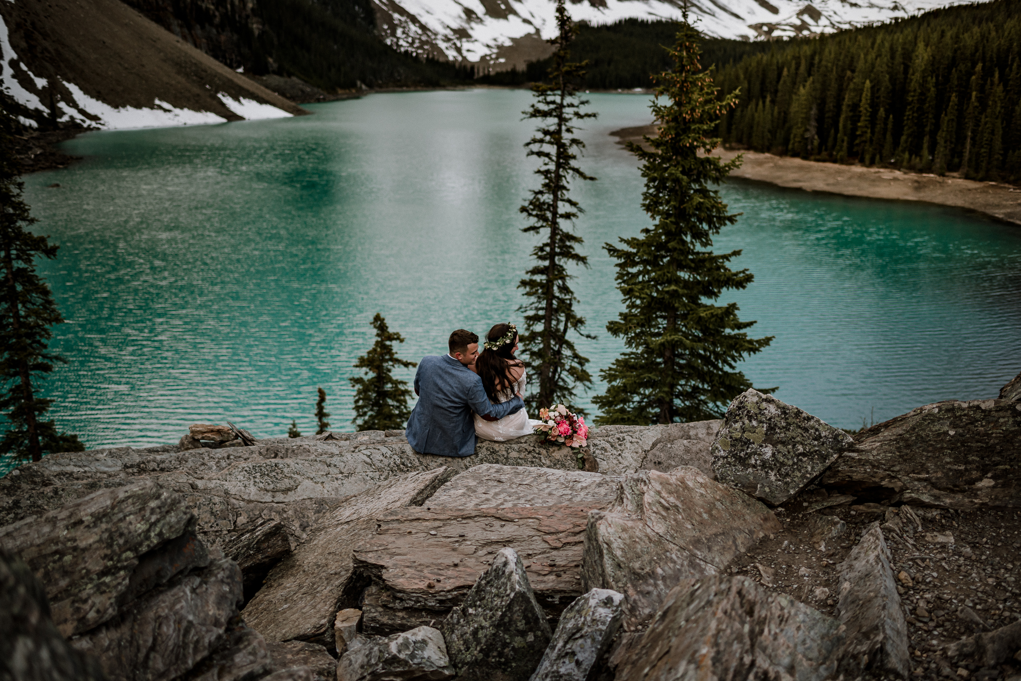 Groom sitting next to bride on a rock at Moraine Lake, Alberta with bouquet beside bride