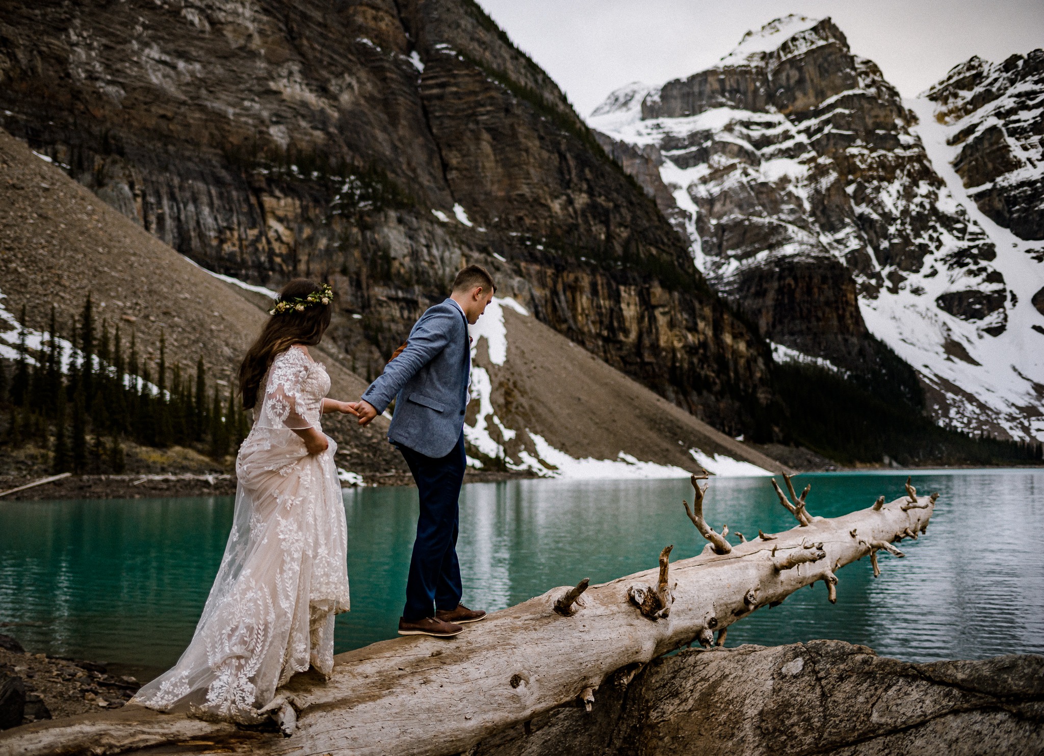 Groom holding brides hand walking up a log at Moraine Lake, Alberta