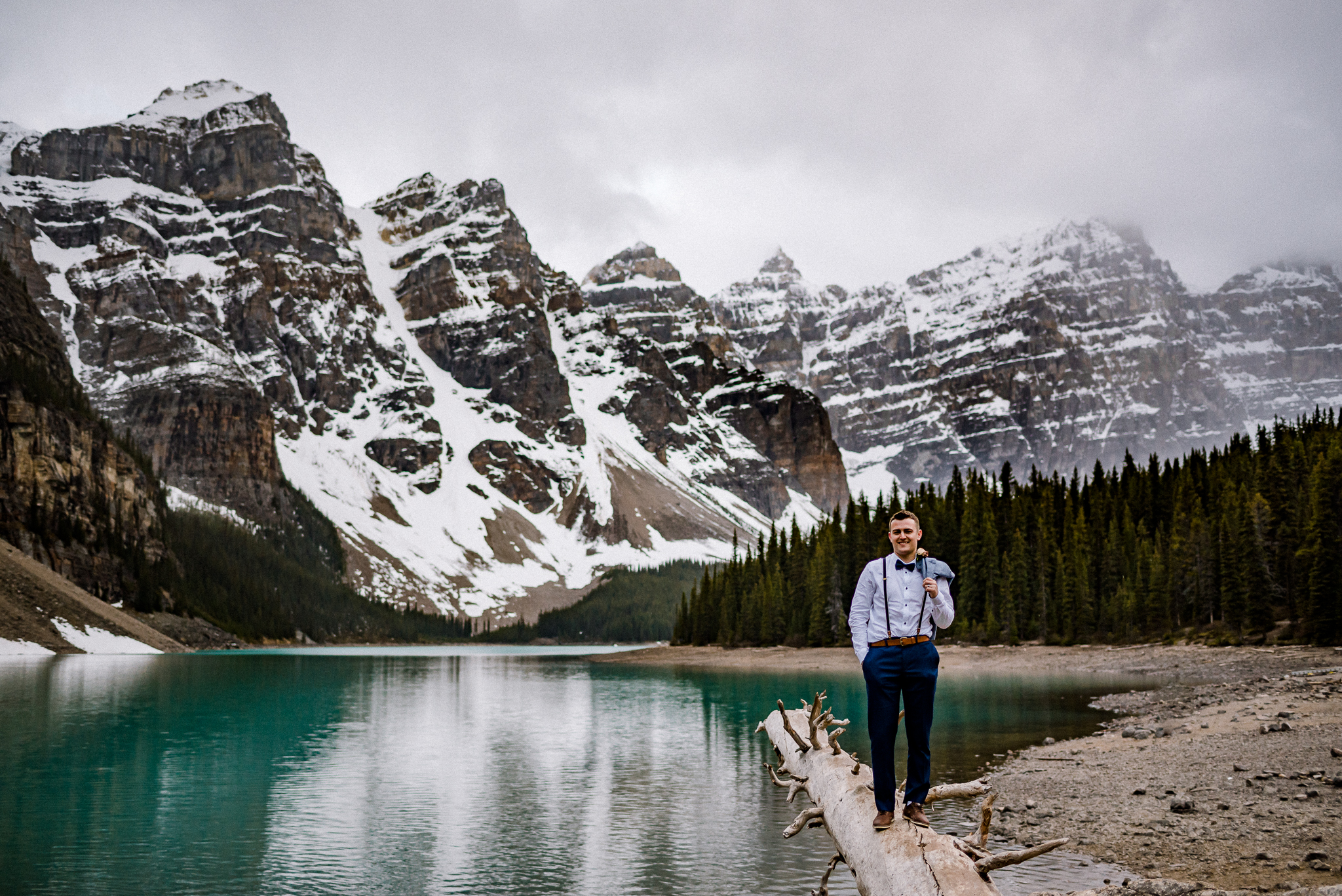 Groom standing on log with jacket slung over shoulder at Moraine Lake, Alberta